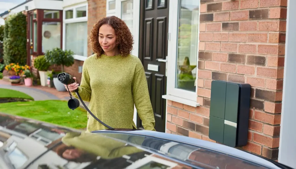A smiling woman with curly brown hair, wearing a lime green sweater, is holding an EV charging cable and standing next to a black electric car and a dark gray home EV charging station mounted on a brick wall.
